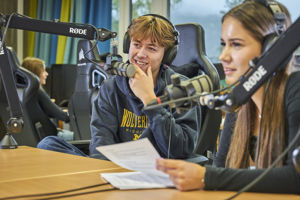 Bildet viser to elevar som sit med mikrofon og manus, og speler inn podcast.