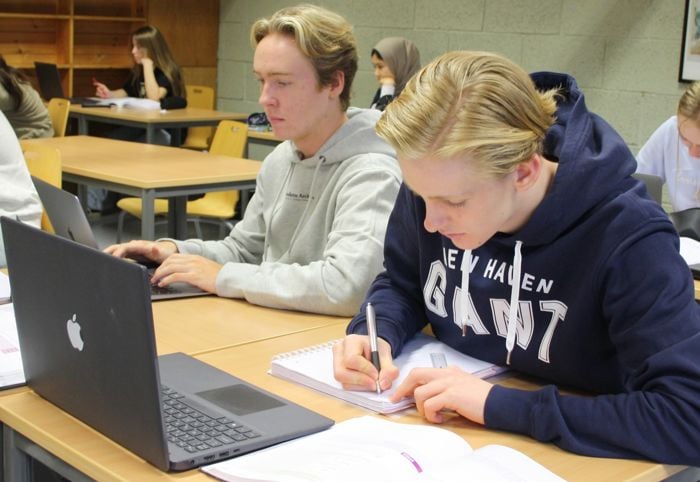Bilde av to gutar som sit ved ein pult i eit klasserom, den eine jobbar på PC og den andre skriv i ei notatbok.
